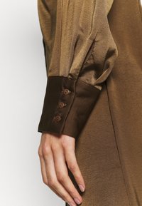 Brązowa satynowa koszula z marszczonymi rękawami, mankietem z trzema guzikami oraz gładką teksturą. Zbliżenie na bok i ramię.