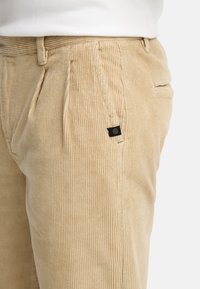 Beige corduroy trousers feature vertical ribbing, front pleats, and a black branded tag on the waistband. Textured, with side pockets.
