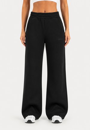 Femme portant un pantalon de survêtement noir à jambes larges avec poches, taille élastique et baskets blanches, debout devant un fond uni.