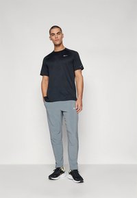 Álló férfi fekete Nike pólóban, világosszürke Nike nadrágban és fekete-fehér Nike sportcipőben, egyszínű világos háttér előtt.
