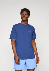 Man in blauwe Patagonia t-shirt en lichtblauwe shorts staat tegen een effen lichte achtergrond en kijkt iets naar de zijkant.