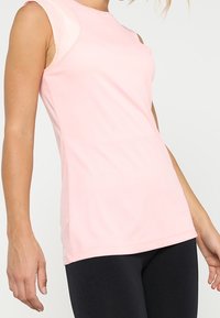Top deportivo sin mangas de color rosa claro, hecho de tela suave, con escote redondo y silueta ajustada, combinado con leggings negros.