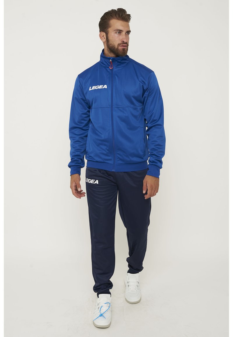 Giacca sportiva blu con colletto alto, chiusura a zip e logo, abbinata a pantaloni blu navy e sneakers bianche con lacci blu.