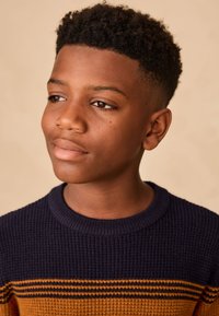 Pull en maille présentant des rayures bleu marine et ambre, avec une finition texturée et un col rond. Gros plan sur le visage d'une jeune personne.
