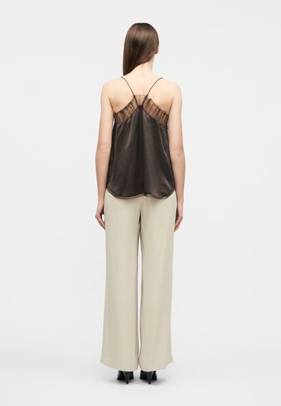 Γυναίκα με μακριά καστανά μαλλιά φοράει μια σκουρόχροη καφέ μεταξωτή καμιζόλα με δαντελένια πλάτη και μπεζ παντελόνια φαρδιά, στέκεται πλάτη προς το φακό μπροστά σε φόντο μονόχρωμο.