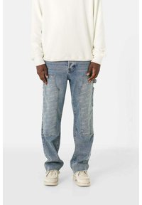 Helle blaue Denim-Jeans in lockerer Passform, mit kontrastierendem Patchwork, frontalen Taschen und ausgefransten Säumen. Dazu wird ein weißer Pullover getragen.