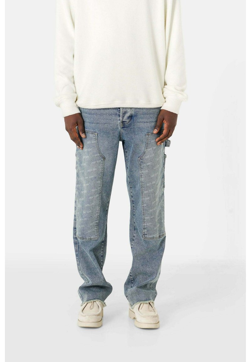 Helle blaue Denim-Jeans in lockerer Passform, mit kontrastierendem Patchwork, frontalen Taschen und ausgefransten Säumen. Dazu wird ein weißer Pullover getragen.
