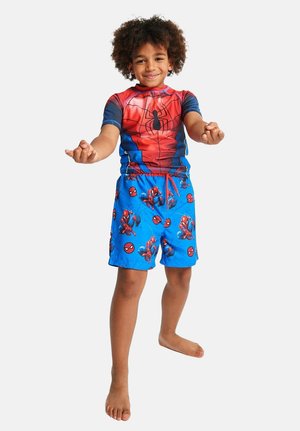 Young child smiling, wearing a Spider-Man costume top and blue shorts with Spider-Man graphics, standing barefoot with hands extended forward.