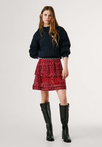 Maglione lavorato a maglia blu navy con maniche a sbuffo, gonna rossa a fantasia e stivali neri in pelle al ginocchio. La gonna presenta un design a strati con una finitura trasparente.