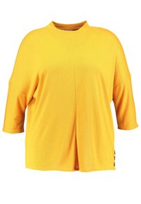 Camiseta oversized amarilla con cuello alto, mangas cortas y detalles de botones en los laterales. Fabricada con una tela suave y texturizada.