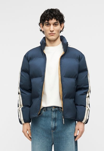 Noor noormehel lokkis juuksed, seljas tumelilla pufferjakk valgete triipudega varruka külgedel, valge särk ja sinised teksad, taustaks hele pind.