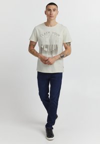 Camiseta gris claro con texto en negrita "RECLAMA TU DENIM" y jeans azul oscuro. El modelo lleva zapatillas negras y tiene tatuajes visibles.