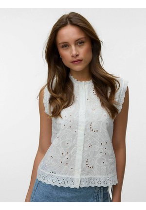 Femme aux longs cheveux bruns portant un chemisier blanc sans manches brodé et un jean en denim bleu sur un fond uni.