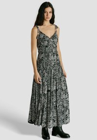 Moteris su ilgais tamsiais plaukais, vilkinti juodai baltos spalvos raštuotą ilgą be rankovių suknelę ir juodus aulinukus, stovinti priešais vienodus foną.