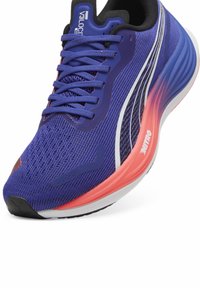 Zapatilla deportiva morada con parte superior de malla, acentos contrastantes en blanco y naranja, y suela acolchada. Presenta una puntera redondeada y cordones texturizados.