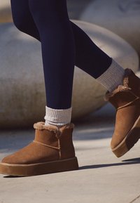 Hnedé semišové členkové topánky s kožušinovým lemovaním, plochými podrážkami a detailmi prešívania, nosené s tmavými legínami a svetlošedými rebrovanými ponožkami.