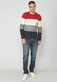 Maglione a righe rosse, crema e blu navy con un design in maglia strutturata, abbinato a jeans in denim blu e sneakers bianche. Presenta un dettaglio del logo.