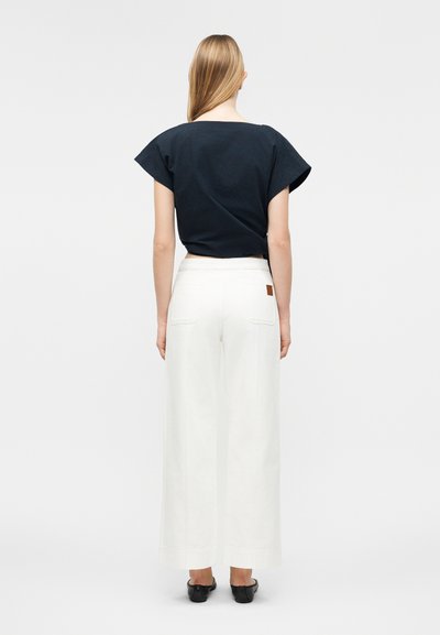 Γυναίκα με μακριά ξανθά μαλλιά φορώντας μαύρο κοντομάνικο τοπ και λευκό παντελόνι ψηλόμεσο με φαρδιά μπατζάκια, στέκεται με την πλάτη προς τα εμπρός.