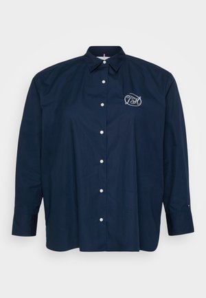 Tummansininen nappipaita, jossa on terävä kaulus, pitkät hihat ja vasemmassa rinnassa valkoinen brodeerattu logo. Sileä pinta.