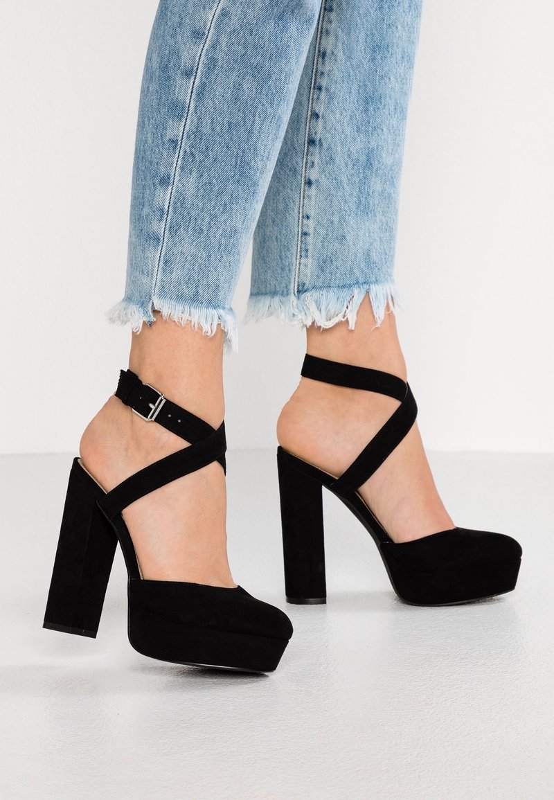 Pieds portant des talons compensés en daim noir avec des brides autour de la cheville, associés à un jean à ourlet effiloché bleu clair, sur fond blanc.
