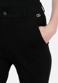 Pantaloni neri con una texture liscia, dotati di chiusura con bottoni, tasche laterali e un dettaglio con logo dorato sul davanti.