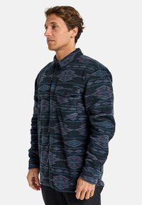 Camicia in pile con una base blu scuro e motivi a diamante viola e grigi, con bottoni davanti e una sola tasca sul petto. Tessuto morbido.