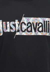 Μαύρο ύφασμα με τη λέξη «justcavalli» με μεγάλα γράμματα, το ένα μισό με floral σχέδιο και το άλλο μισό μονόχρωμο μαύρο σε λευκό ορθογώνιο φόντο.
