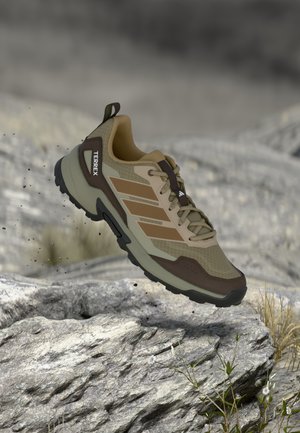Chaussure de trail marron clair avec accents gris, dessus en mesh, surface texturée et semelle robuste. Présente le logo "TERREX" et un embout renforcé.