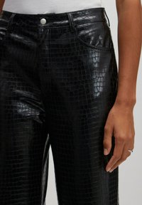 Schwarze Kunstlederhosen mit einem krokodilgeprägten Muster, mittelhoher Taille, vorderen Taschen und einem silbernen Knopfverschluss.