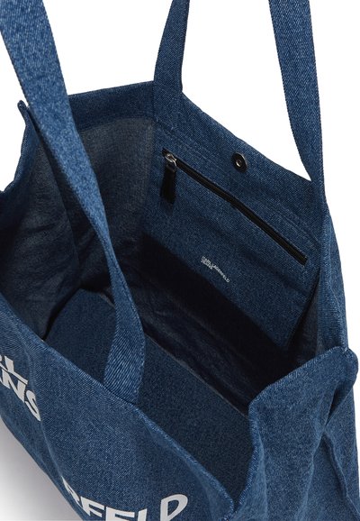 Sac fourre-tout en denim avec de longues poignées, de couleur bleu profond, poche intérieure zippée et logo imprimé sur le côté. Tissu texturé.