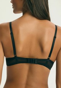 Žena zobrazená zozadu v čiernej podprsenke s nastaviteľnými ramienkami a zapínaním na háčiky na svetlom pozadí.