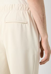 Pantalones beige con una tela texturizada, que cuentan con una cinturilla elástica y un solo bolsillo trasero con una sutil etiqueta de logo.