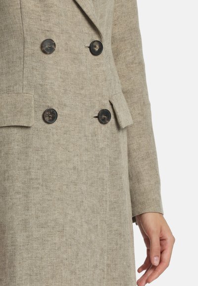 Gros plan sur un manteau en lin beige à double boutonnage avec des boutons foncés et des poches à rabat, montrant la main droite d'une personne sur le côté.