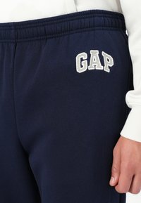 Mørkeblå joggebukser laget av mykt stoff, med et brodert hvitt "GAP"-logo på venstre lår og elastisk midje.