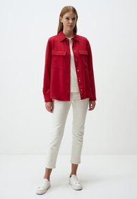 Chemise rouge à boutons avec deux poches poitrine, manches longues et coupe décontractée, associée à un pantalon blanc et des baskets blanches.