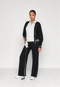 Μαύρο πλεκτό φόρμα με ζακέτα με φερμουάρ και παντελόνια με φαρδιά μπατζάκια. Λευκό πουκάμισο με γιακά από μέσα. Το μοντέλο κρατάει ένα γκολφ πατερίτσα και φοράει λευκό γάντι.