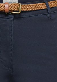 Pantalon bleu marine avec une ceinture en cuir tressé couleur fauve passée dans les passants, montrant les coutures et une étiquette partiellement visible à l'intérieur.