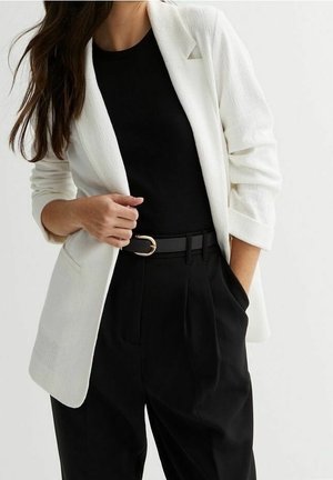 Femme portant un blazer blanc sur un haut noir, un pantalon noir taille haute avec une ceinture noire ornée d'une boucle dorée, posant avec une main dans la poche.