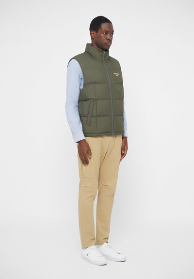 Nabran oljčnozeleni jopič z visokim ovratnikom, sprednjim zadrgo in stranskimi žepi, nosi se čez svetlo modro srajco in bež hlače.