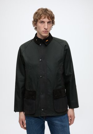 Junger Mann mit hellem lockigem Haar, der eine dunkelgrüne Jacke mit schwarzem Kragen und Taschen trägt, steht vor einem schlichten weißen Hintergrund.