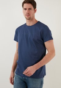 Tamsiai mėlyna medvilninė marškinėlių suapvalintu kaklu, trumpomis rankovėmis ir subtiliu iškilmingu tekstu priekyje. Glotni tekstūra, laisvas kirpimas.