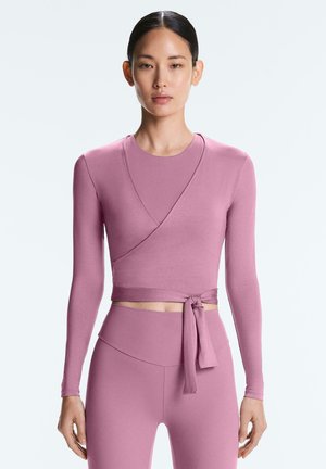 Frau trägt langärmliges Wickel-Crop-Top und hoch taillierte Leggings, beide in staubigem Rosa, stehend vor einem schlichten hellen Hintergrund.