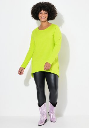 Neon-grünes Langarmoberteil mit rundem Ausschnitt, kombiniert mit schwarzen Lederleggings und glänzenden lilafarbenen Cowboystiefeln, vor einem weißen Hintergrund stehend.