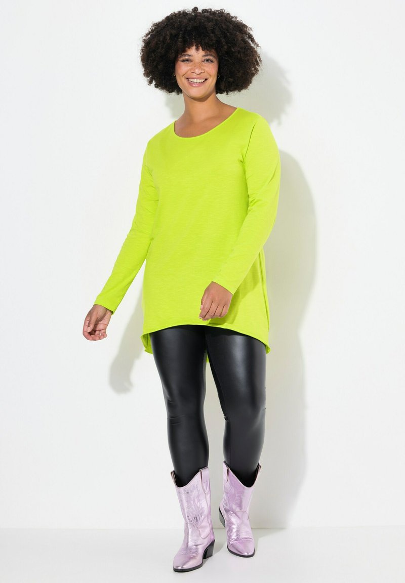Neon-grünes Langarmoberteil mit rundem Ausschnitt, kombiniert mit schwarzen Lederleggings und glänzenden lilafarbenen Cowboystiefeln, vor einem weißen Hintergrund stehend.