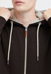 Bruine zip-up hoodie met een gezellig crème fleece binnenkant, koordjes en een leren label. Heeft een gladde textuur en zilveren hardware.