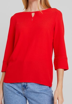 Blouse rouge légère à manches trois-quarts, avec un décolleté en trou de serrure et des détails en dentelle sur les manches. Associée à un jean bleu.