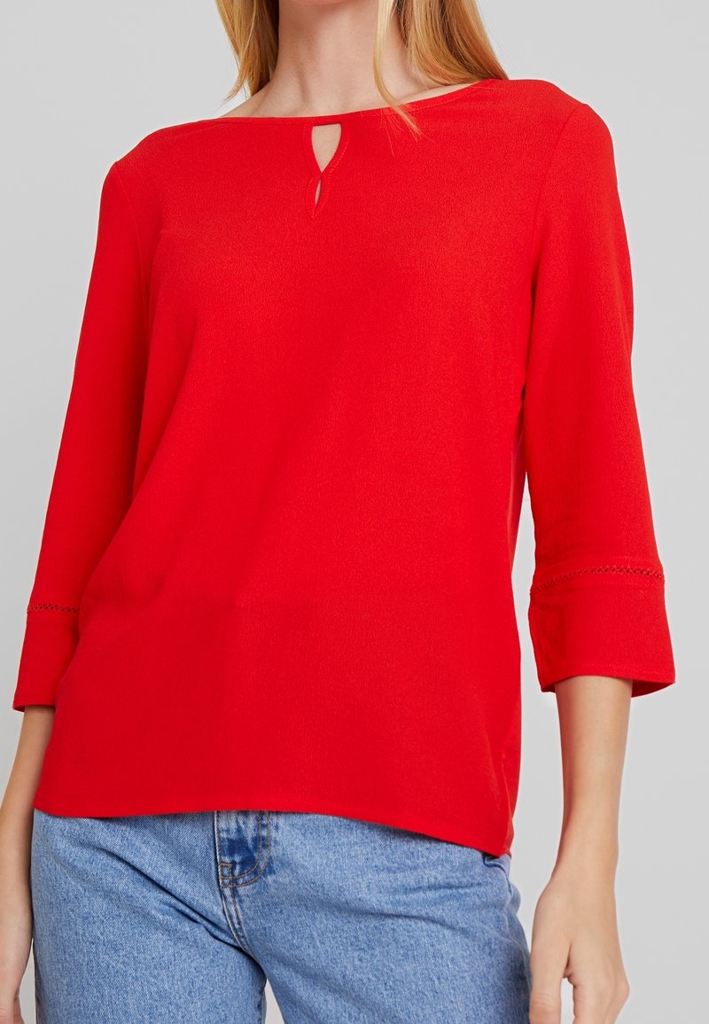 Blouse rouge légère à manches trois-quarts, avec un décolleté en trou de serrure et des détails en dentelle sur les manches. Associée à un jean bleu.