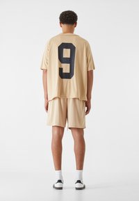 Ensemble de maillot et de shorts de sport beige en tissu mesh ; le maillot présente un grand numéro noir 9 au dos. Chaussettes et chaussures blanches.