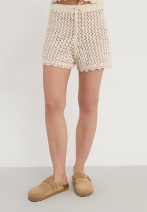 Persona que lleva puestos unos pantalones cortos de punto crochet crema con bordes en forma de concha y zuecos marrones sin cordones, de pie sobre un suelo neutro.