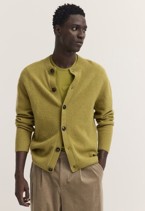 Man wearing a mustard yellow cardigan over a matching t-shirt and beige corduroy pants, looking to his left against a plain background.